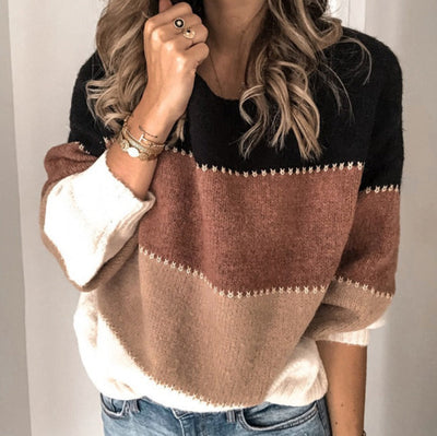 Femme portant un pull tricolore noir, marron et beige, manches longues, avec des bijoux dorés, sur fond intérieur neutre. Mode automne-hiver tendance.