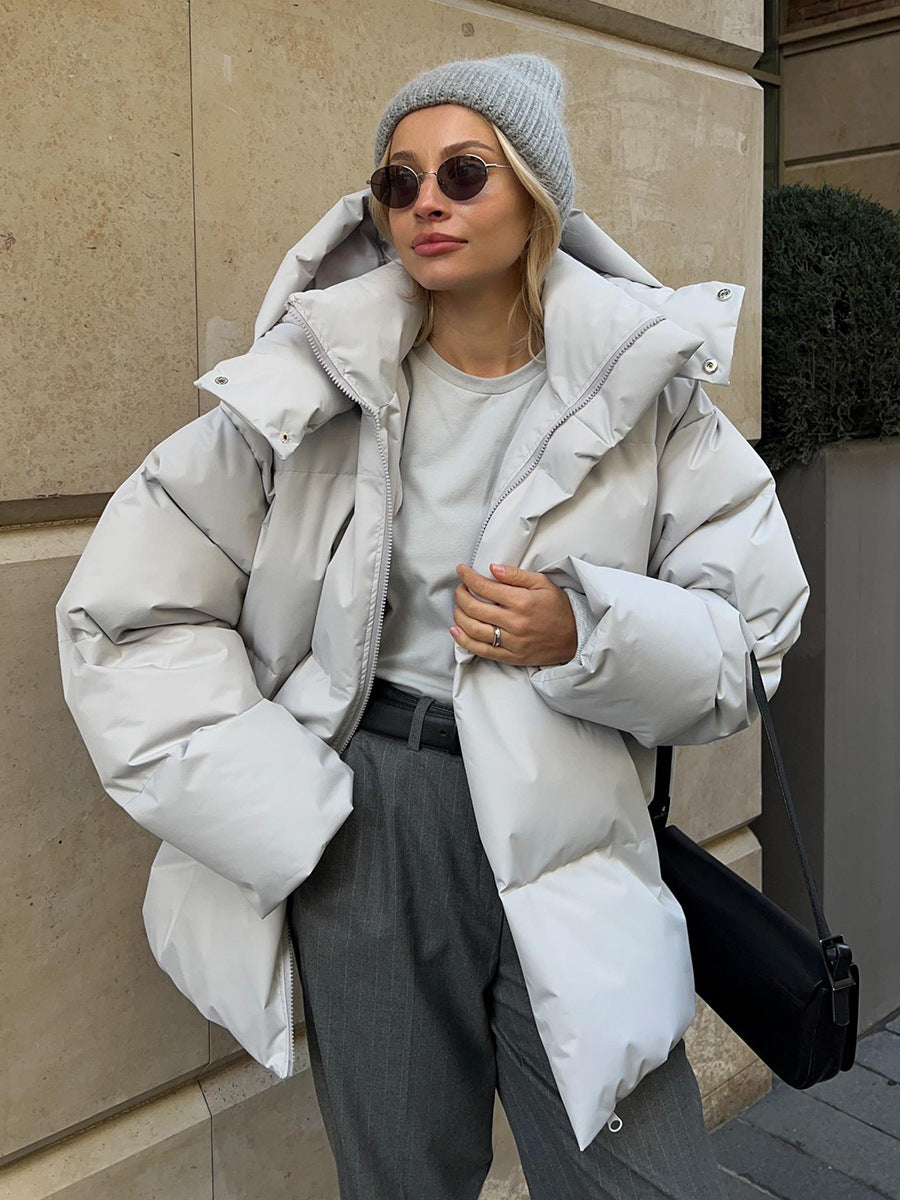 Femme portant une doudoune blanche, bonnet gris et lunettes de soleil, posant contre un mur en pierre. Mode hiver chic, style urbain élégant.