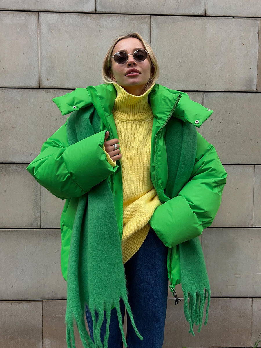 Femme en manteau vert vif, pull jaune, écharpe verte, lunettes de soleil rondes, posant devant un mur en pierre. Mode hiver tendance, style urbain.