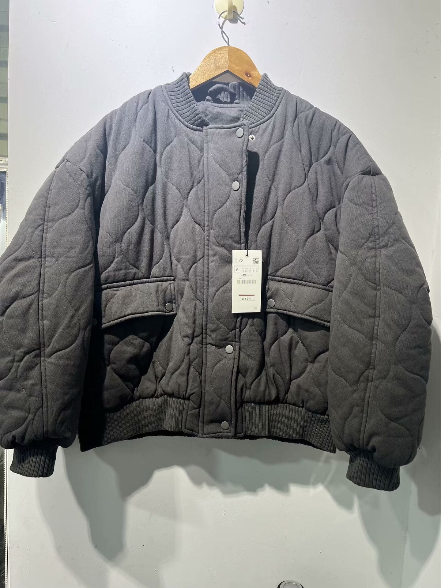 Veste matelassée grise sur cintre, design moderne, col rond, poches avant, étiquette visible. Mode hiver, tendance, vêtements chauds.