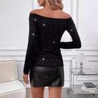 Femme portant un haut noir à paillettes et une jupe en cuir noir, debout dans un salon moderne avec canapé blanc et table en verre.