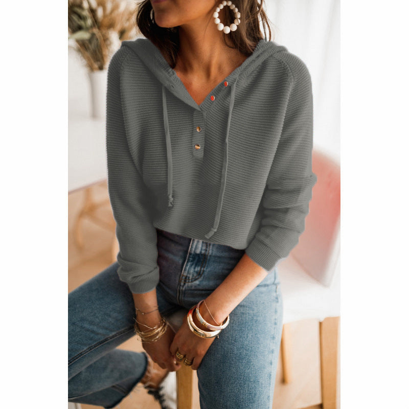Femme portant un pull à capuche gris avec boutons, assise sur une chaise. Style décontracté, jeans, boucles d'oreilles pendantes, bracelets dorés.