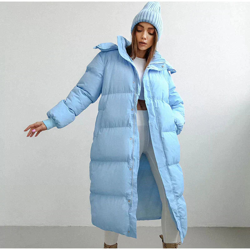 Femme portant un manteau long matelassé bleu clair et un bonnet assorti, posant dans un intérieur minimaliste. Mode hiver, style décontracté.