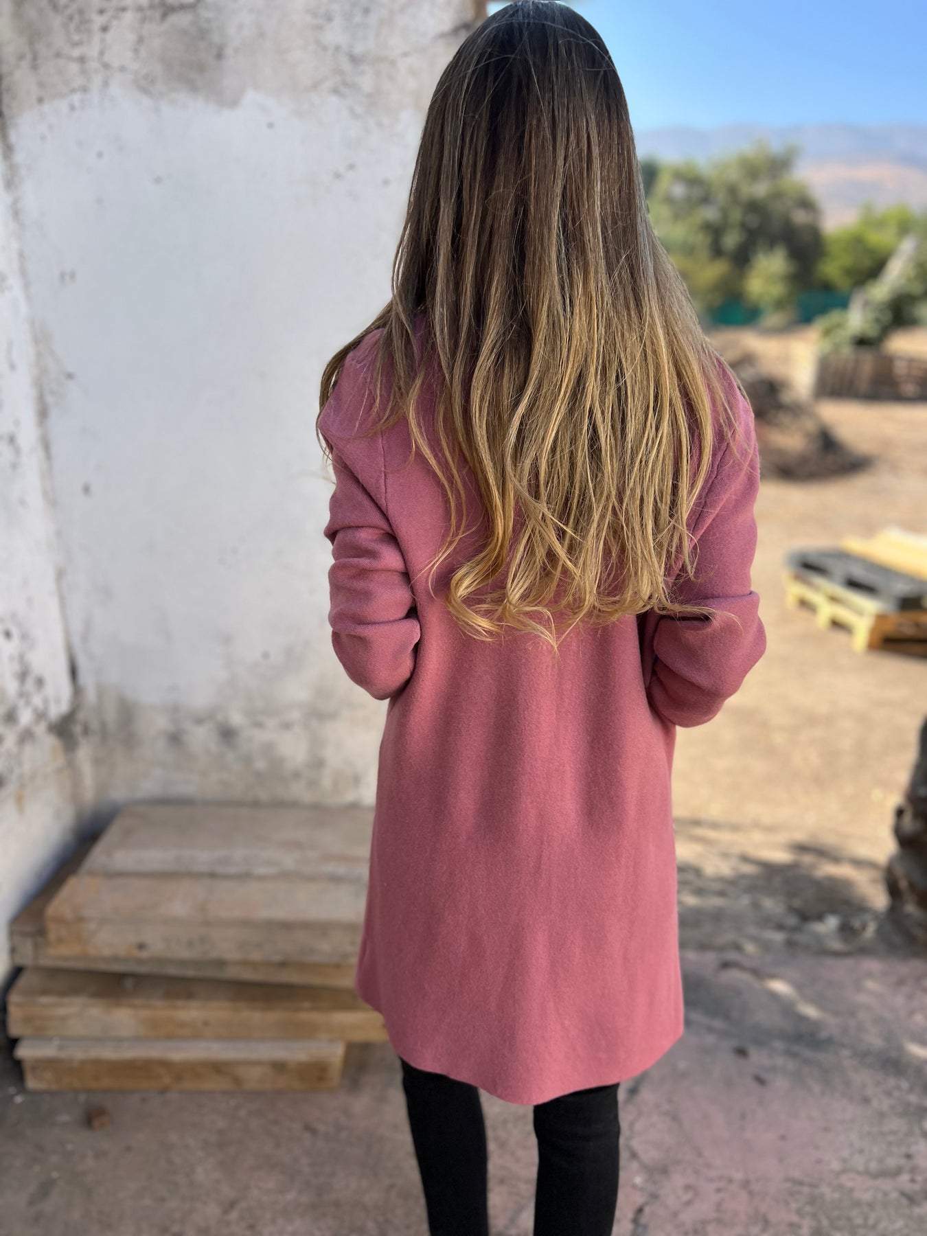 Femme de dos avec longs cheveux blonds, portant un manteau rose, debout à l'extérieur près de palettes en bois, paysage flou en arrière-plan.