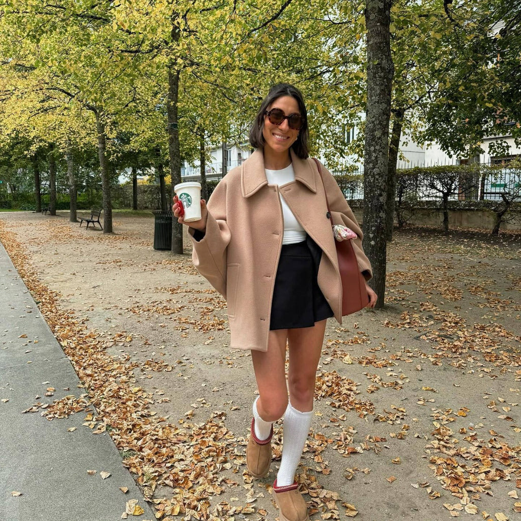 Jeune femme souriante en manteau beige, jupe noire et chaussettes hautes blanches, tenant un café Starbucks, marchant dans un parc automnal.