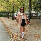 Jeune femme souriante en manteau beige, jupe noire et chaussettes hautes blanches, tenant un café Starbucks, marchant dans un parc automnal.