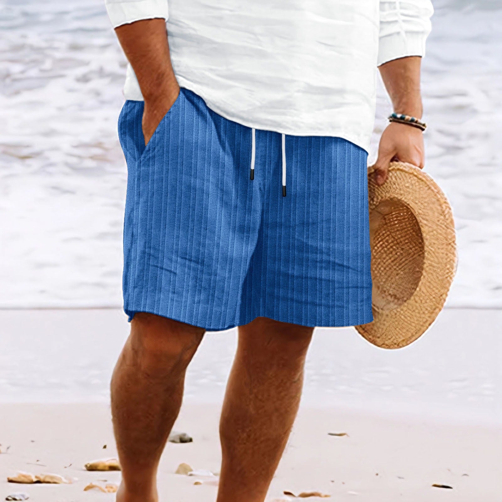 Homme en short bleu rayé, tenant un chapeau de paille sur une plage. Mode estivale, tenue décontractée, bord de mer.