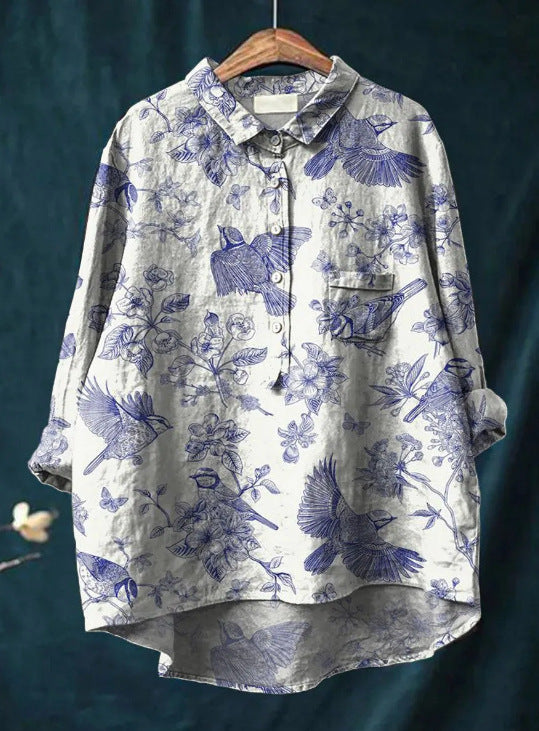 Chemise ample en lin gris avec motifs bleus d'oiseaux et fleurs, manches longues, col boutonné, suspendue sur un cintre en bois.