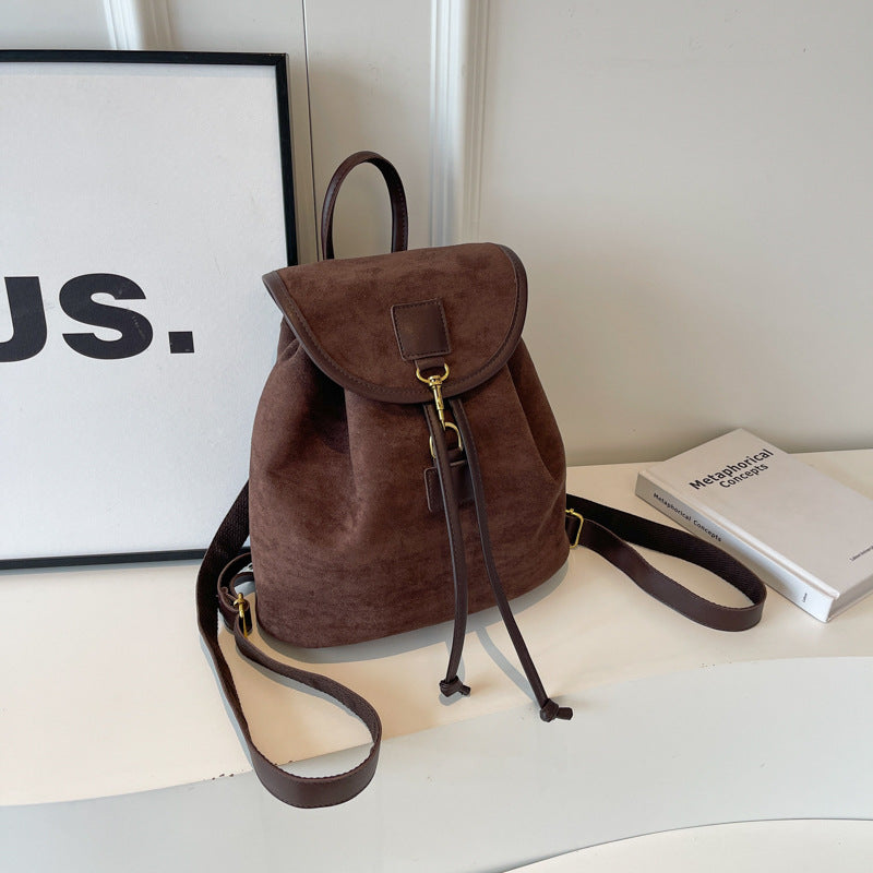 Sac à dos en daim marron avec cordons et boucle dorée, posé sur une table blanche à côté d'un livre et d'un cadre minimaliste.