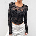 Femme portant un haut transparent noir à motifs floraux, manches longues, associé à un pantalon blanc. Mode élégante et tendance.