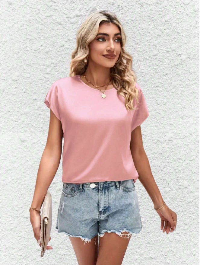 Femme blonde portant un t-shirt rose et un short en jean déchiré, tenant une pochette beige, posant devant un mur blanc texturé. Mode estivale décontractée.