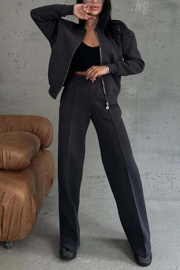 Femme en ensemble de survêtement noir, veste zippée et pantalon ample, posant devant un mur gris. Mode décontractée, tendance automne-hiver.