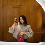 Femme prenant un selfie dans un miroir, portant une veste grise et tenant un sac à main rouge. Fond en bois, ambiance élégante et moderne.