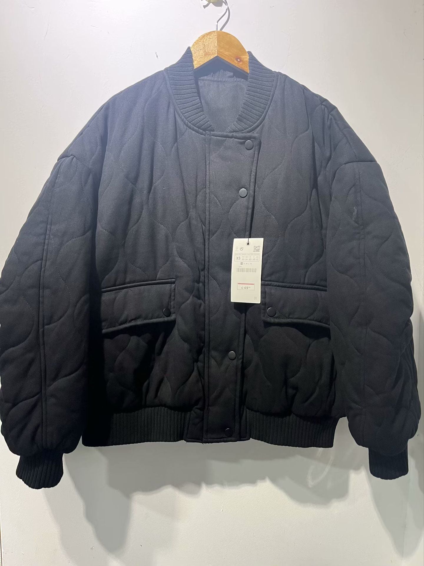 Veste matelassée noire sur cintre en bois, avec étiquette. Design moderne, col rond, poches avant. Mode hiver, tendance vestimentaire.