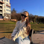 Jeune femme assise en extérieur, portant des lunettes de soleil noires et une veste beige, devant un bâtiment moderne et un parc ensoleillé.