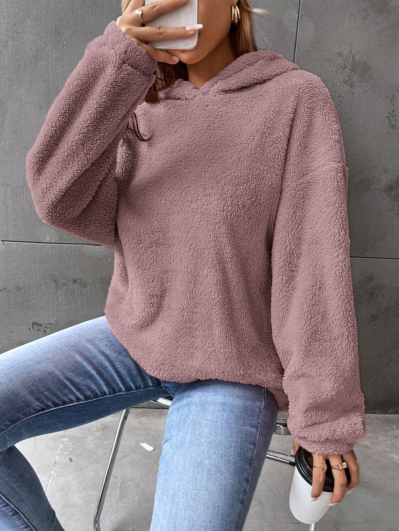 Femme portant un pull en peluche rose, tenant un smartphone et un gobelet, assise sur une chaise. Mode décontractée, tendance automne-hiver.