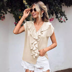 Femme souriante portant un haut beige à volants et un short blanc déchiré, lunettes de soleil, posant devant un mur blanc avec des fleurs.