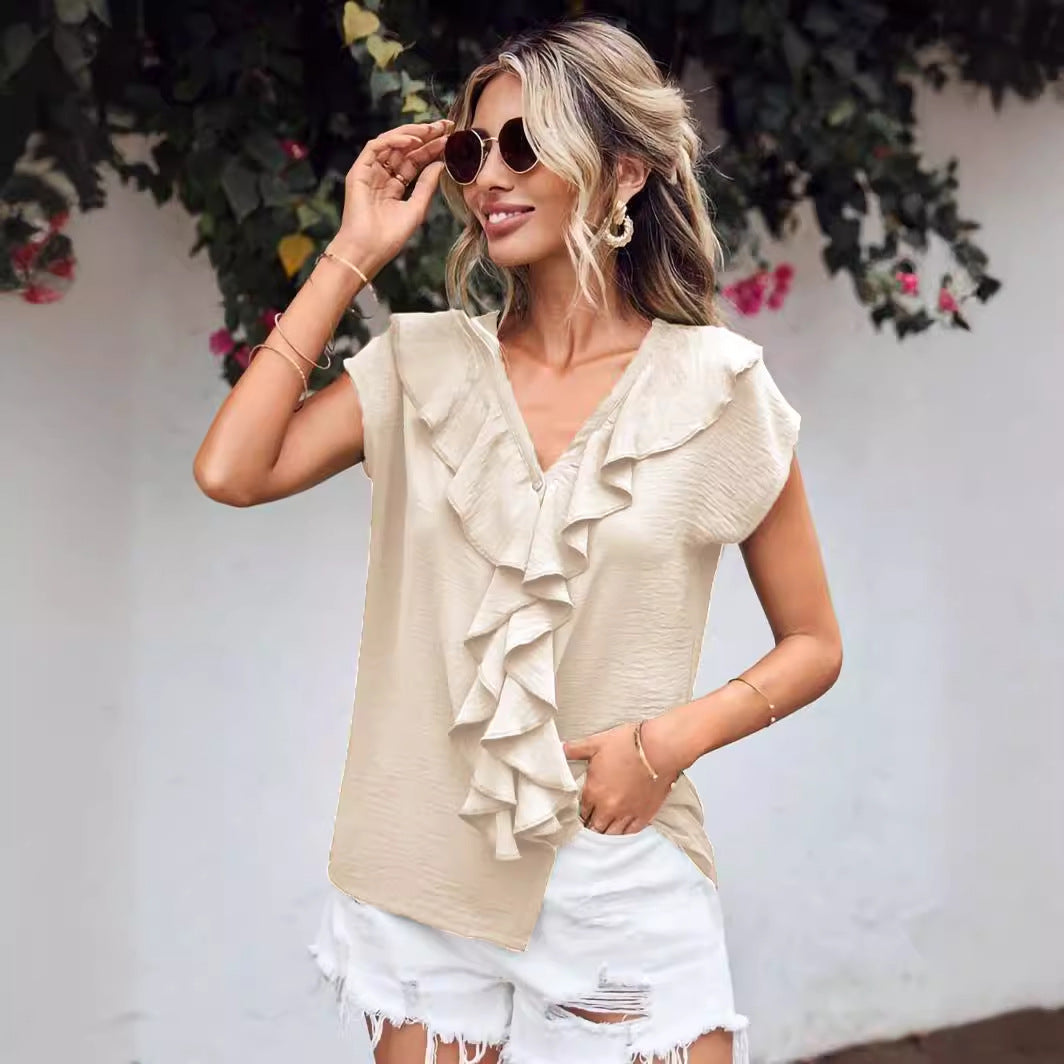 Femme souriante portant un haut beige à volants et un short blanc déchiré, lunettes de soleil, posant devant un mur blanc avec des fleurs.