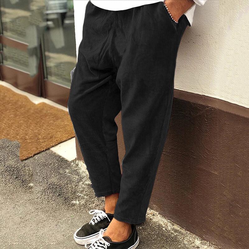 Pantalon noir ample pour homme, style décontracté, porté avec des baskets noires et blanches, devant un mur beige et une porte vitrée.