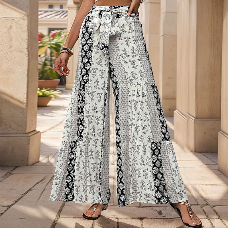 Pantalon large à motifs floraux noir et blanc, ceinture nouée, style bohème chic, femme en sandales, fond architectural élégant.