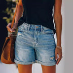 Femme portant un short en jean délavé avec un haut noir, tenant un sac en cuir marron. Mode estivale décontractée, tendance et élégante.