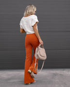Femme de dos portant un pull blanc et un pantalon orange évasé, tenant un sac beige devant une porte de garage grise. Mode automne chic.