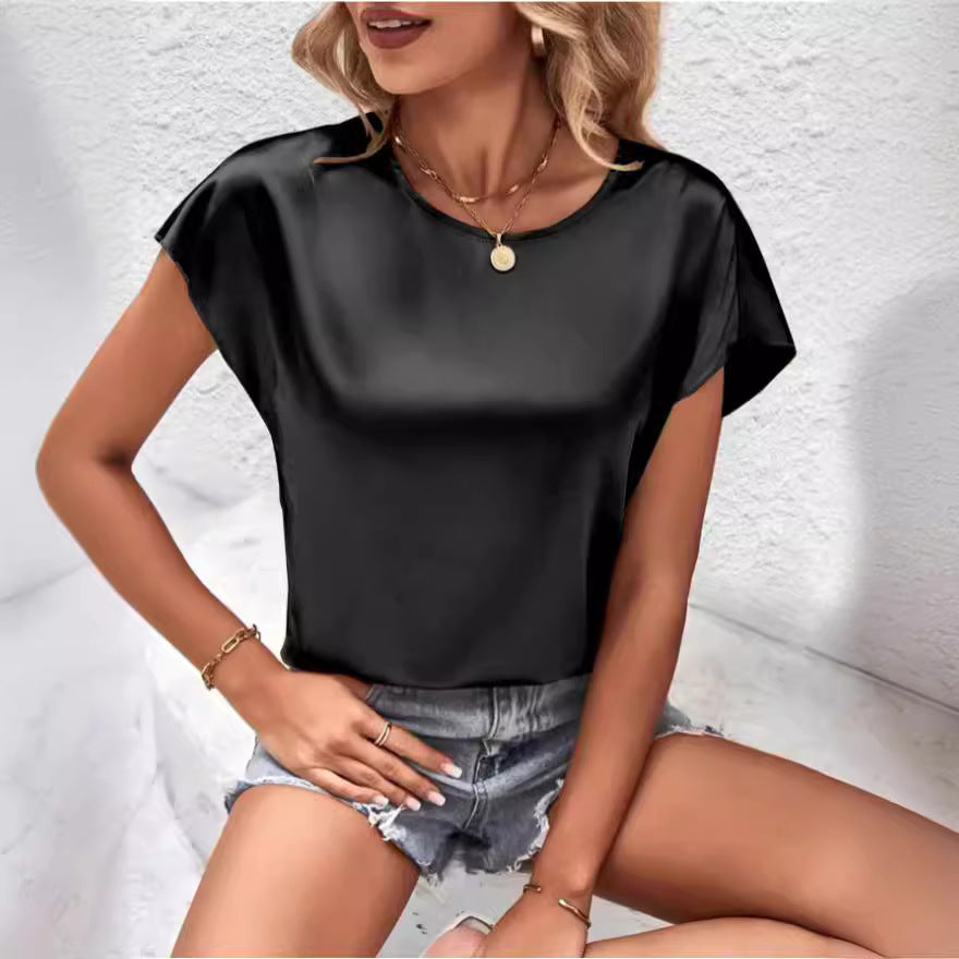 Femme portant un haut en satin noir et un short en jean déchiré, assise contre un mur blanc. Mode estivale chic, tenue décontractée élégante.