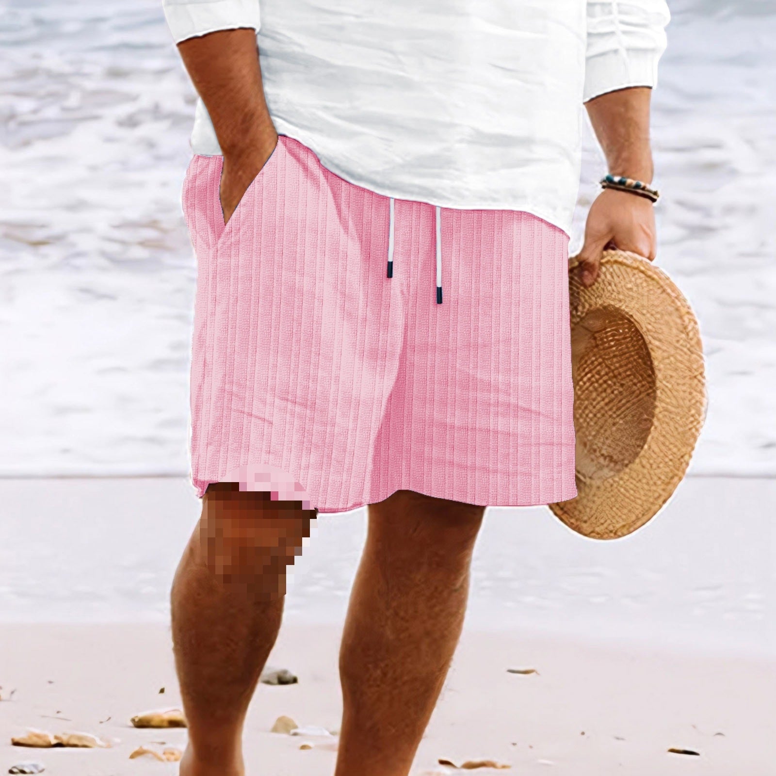 Homme en short rose rayé, tenant un chapeau de paille, debout sur une plage. Mode estivale, tenue décontractée, bord de mer.
