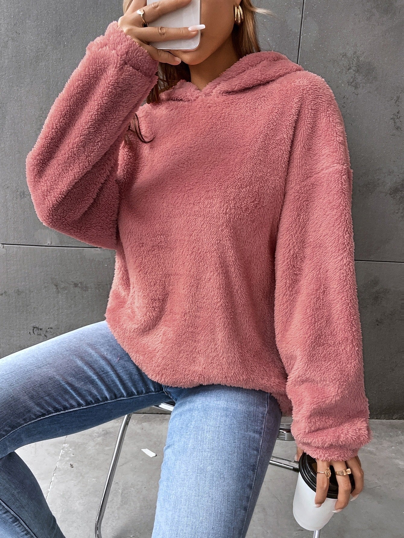 Femme portant un pull en peluche rose, assise sur une chaise, tenant un smartphone et un gobelet. Mode décontractée, tendance automne-hiver.