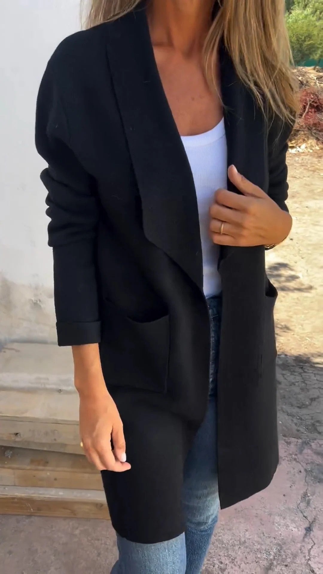 Femme portant un long manteau noir élégant, débardeur blanc et jean, posant à l'extérieur par temps ensoleillé. Mode automne chic et décontractée.