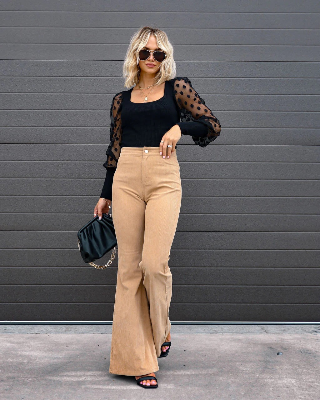 Femme blonde portant un haut noir à manches transparentes et un pantalon beige évasé, lunettes de soleil, sac noir, posant devant un mur gris.