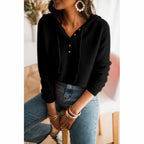 Femme portant un pull noir à capuche avec boutons, jeans décontracté, assise sur une chaise, accessoires élégants, boucles d'oreilles pendantes.