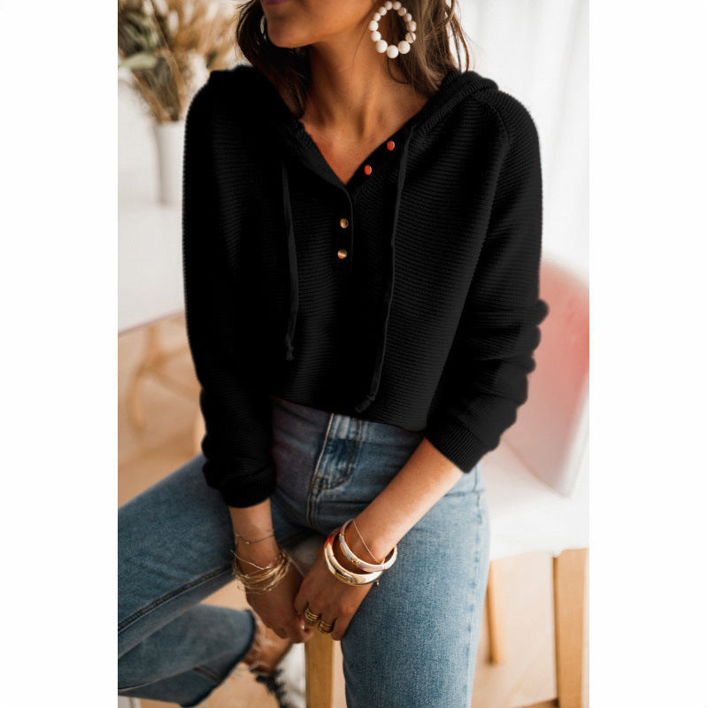Femme portant un pull noir à capuche avec boutons, jeans décontracté, assise sur une chaise, accessoires élégants, boucles d'oreilles pendantes.