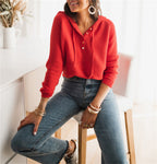 Femme portant un pull rouge à capuche et un jean taille haute, assise sur un tabouret dans un intérieur lumineux. Mode décontractée, style moderne.