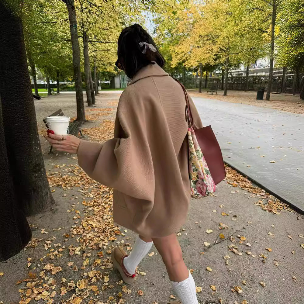 Femme en manteau beige marchant dans un parc automnal, tenant un café à emporter, sac à main fleuri, feuilles mortes au sol, ambiance décontractée.