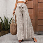 Femme portant un pantalon large à motifs floraux beige, tenant un sac en osier, devant une porte en bois, plantes vertes en arrière-plan.