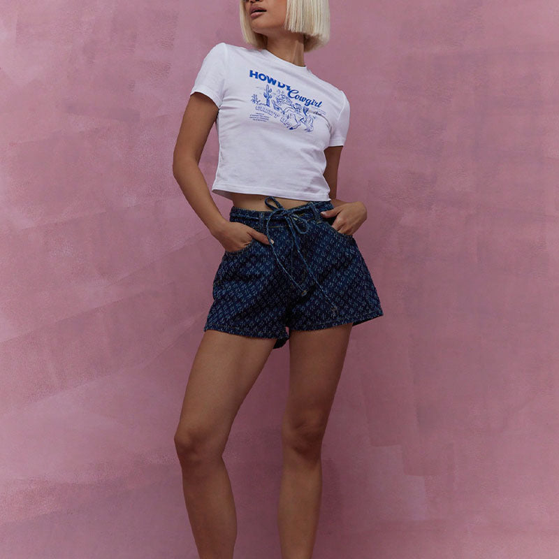 Femme en t-shirt blanc et short bleu, posant devant un mur rose. Mode estivale, tenue décontractée, style moderne.