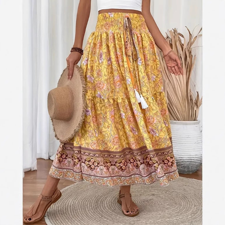 Jupe bohème longue jaune à motifs floraux, ceinture élastique, tenue estivale décontractée, femme tenant un chapeau de paille, ambiance chic et naturelle.