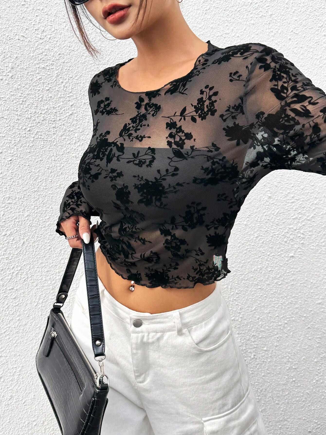 Femme portant un haut transparent noir à motifs floraux, tenant un sac à main noir texturé, pantalon blanc; mode élégante et tendance.