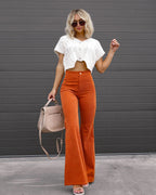 Femme en pull blanc court et pantalon orange évasé, portant un sac beige, posant devant un mur gris. Mode automne, style chic et décontracté.