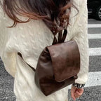 Femme portant un sac à dos en cuir marron sur un pull blanc, traversant une rue. Mode automne, accessoires tendance, style urbain chic.