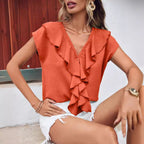 Femme portant un chemisier orange à volants, assise, avec des boucles d'oreilles dorées et un short blanc déchiré. Mode estivale élégante.