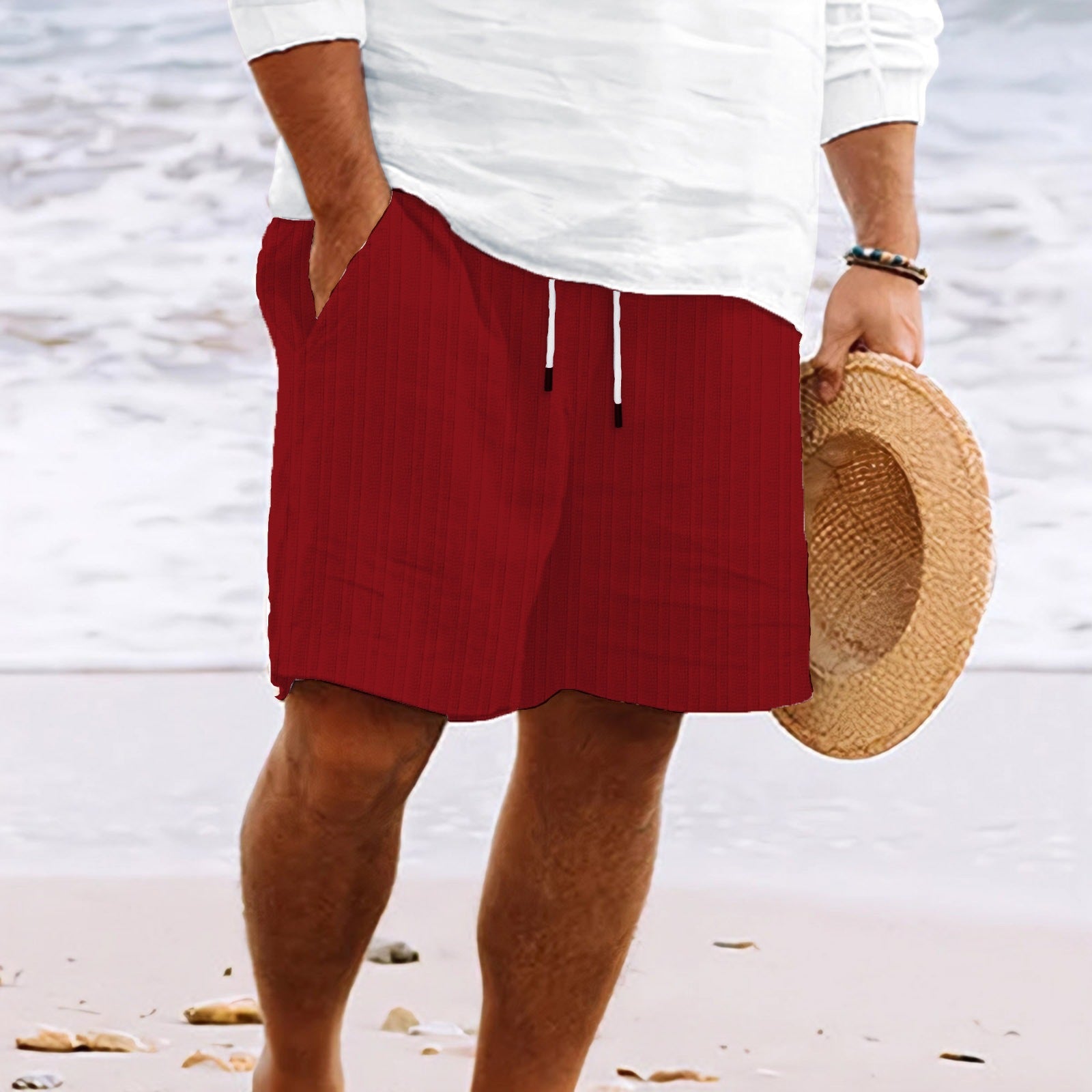 Homme en short rouge et chemise blanche tenant un chapeau de paille sur une plage. Mode estivale, tenue décontractée, vacances bord de mer.