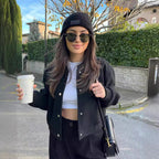 Jeune femme souriante portant une veste noire, un bonnet et des lunettes de soleil, tenant un café à emporter dans une rue bordée de verdure.