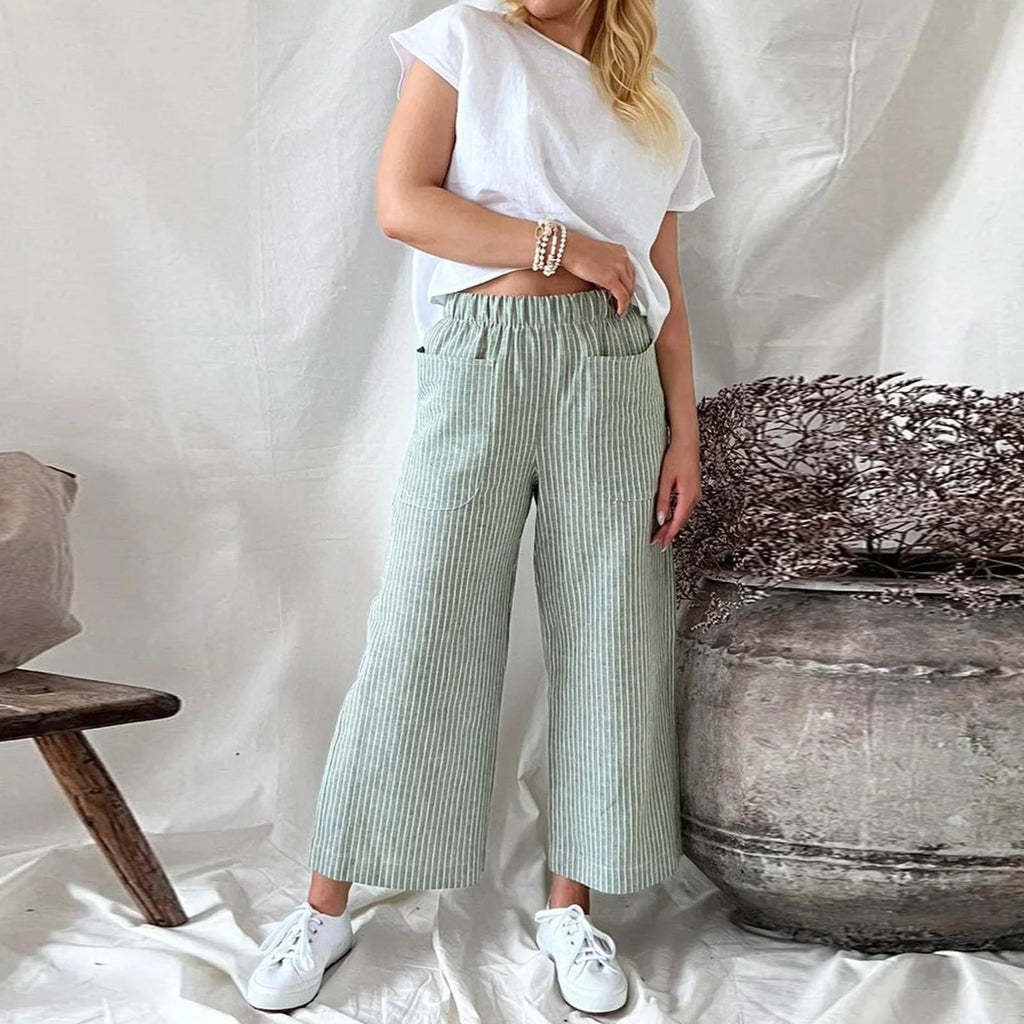 Femme en pantalon large rayé vert, t-shirt blanc, posant près d'un pot en céramique, style décontracté, mode printemps-été, intérieur minimaliste.