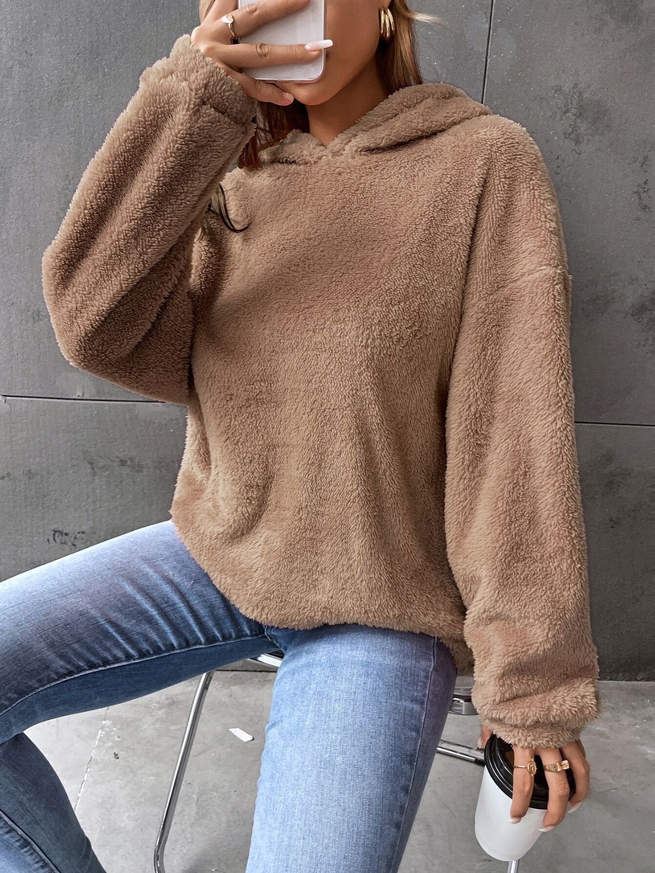 Femme portant un pull en peluche beige, tenant une tasse de café, assise sur une chaise. Mode décontractée, tendance automne, confort.