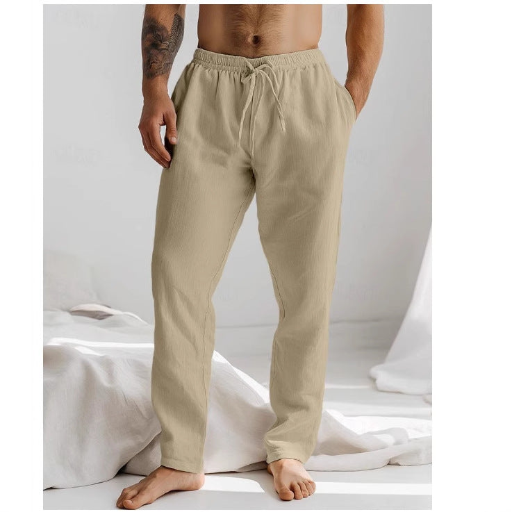 Homme portant un pantalon de détente beige en lin, pieds nus, debout dans une chambre lumineuse. Mode masculine, confort, style décontracté.