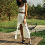 Femme en ensemble blanc côtelé, jupe fendue, pose sur terrasse en bois, fond de nature verdoyante, mode estivale élégante, sandales noires.