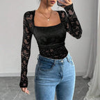 Femme portant un haut en dentelle noire à manches longues et un jean bleu, posant devant un mur blanc. Mode féminine élégante et moderne.