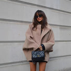 Femme portant un manteau beige oversize, lunettes de soleil noires, et sac à main en cuir noir devant un mur gris. Mode automne chic et tendance.